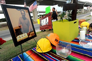 A shrine of hard hats, candles, and a photo of José Lainez, a fallen construction worker, sits on a table at a July 11 vigil near the U.S. 183 construction site where Lainez died June 15. The Workers Defense Project, which sponsored the vigil, says the father of five died of heat exhaustion because adequate water and rest breaks were not provided at the job site.
