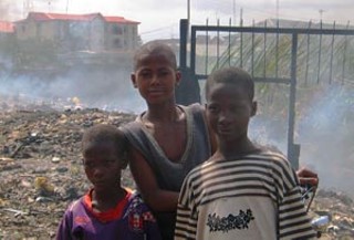 Children standing in front of a smoldering electronic waste dump just outside of the Alaba market in Lagos, Nigeria. Burned electronic waste produces polycyclic aromatic hydrocarbons, dioxins, and heavy metal emissions – carcinogenic and highly toxic chemicals. These children live next to the dump.

<br>
Photo © Basel Action Netwrok 2005